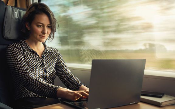 Frau sitzt im Zug und ist auf einen Laptop fokussiert.